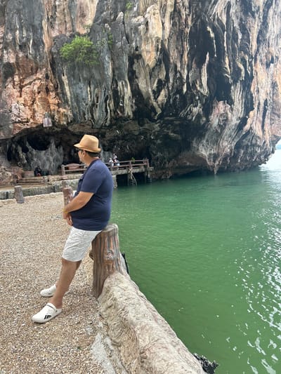 A man wearing a navy blue t-shirt, white patterned shorts, a straw fedora, and white slip-on shoes sits on a log-shaped bench overlooking a body of emerald green water. He is facing to the right, looking out at the water and the massive rock formations that rise dramatically from the sea. The rock face is textured with various shades of gray, brown, and orange, with patches of green vegetation clinging to its surface. In the background, there is a wooden pier and a cave entrance, with a few other people visible in the distance. The ground in the foreground is covered with small, light-colored pebbles. The scene appears to be a scenic viewpoint in Tambon Krasom, Thailand, known for its karst topography and sea caves.