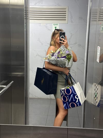 A young woman is taking a mirror selfie in an elevator. She is holding a bouquet of purple and white flowers and two shopping bags. One bag is dark blue with a gold logo, and the other is white with blue and pink lettering. She is wearing a light green ribbed dress and sunglasses on her head. The elevator is modern with stainless steel walls and a gray tiled floor. A sign in Hebrew is visible on the wall. The location is identified as Ariel, Palestinian Territories.