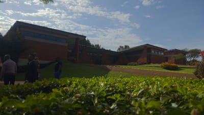 A low-angle shot shows a red brick building complex with a unique, angular roofline against a partly cloudy sky. Lush green grass slopes towards the structure. In the foreground, a hedge with small flowers dominates the view. Several people are indistinctly visible on the left side of the image, walking or standing. A small, secondary building is visible on the right side of the image.