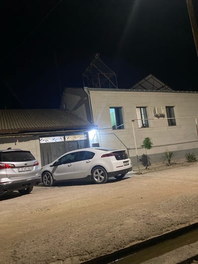 The image depicts a nighttime scene in Verin Artashat, Armenia. Two cars are parked on a dirt road in front of a residential building. The building has a corrugated metal roof and a grey facade with several windows. An air conditioning unit is visible above one of the windows. A silver Chevrolet Equinox is parked on the left, and a white Chevrolet Volt is parked on the right. The license plate of the Chevrolet Equinox reads "37 RR 888". The license plate of the Chevrolet Volt reads "AY 135 AOA". A solar water heater is mounted on the roof of the building, supported by a metal frame. The ground in front of the building is unpaved and appears to be dirt or gravel. A narrow, dark ditch runs along the edge of the road in the foreground. The sky is dark, indicating it is nighttime.