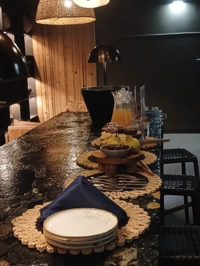 Here is a description of the image:
The image shows a food and beverage service area. The scene appears to be indoors. A table displaying a variety of food and beverage items is featured. There are carafes with liquid in them. Adjacent to the carafes are wooden bowls filled with food items. Plates covered by blue napkins are stacked on top of straw-like placemats. The table is set with flatware. The background has a light fixture and the presence of a wooden slat wall. There are black and grey objects. Due to the limited information from the single image, further details about the location in Nwanaeanoka, Botswana, or specifics about people and actions are unavailable.