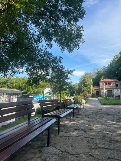 The image depicts a picturesque view of the Madara National Historical-Archaeological Reserve in Madara, Bulgaria. In the foreground, several dark wooden park benches with metal legs are arranged along a cracked stone pathway. The pathway leads towards a building with a red-tiled roof, constructed from stone. A flagpole with the Bulgarian flag stands to the left of the building. Lush green trees and vegetation surround the area, creating a serene and natural ambiance. In the background, there are structures that appear to be kiosks or small shops, and a bright blue truck with an "Oreo" logo. The sky above is blue with scattered white clouds. The scene suggests a peaceful, outdoor location that is likely a tourist attraction.