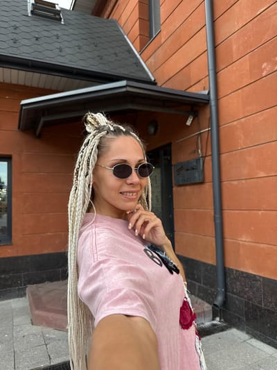In Arkhangel'sk, Russia, a woman with blonde braids and sunglasses is standing in front of a brick building. She is wearing a pink t-shirt with patches and is holding her hand to her chin. The building has a dark grey roof with a skylight and an overhang. A grey downspout runs along the side of the building. The ground is paved with grey tiles.