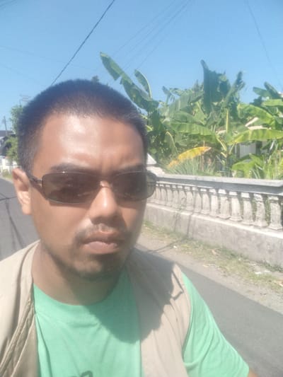 This is a selfie of a man with short, dark hair, wearing dark sunglasses and a light brown vest over a light green t-shirt. He has a mustache and a goatee. The man is outdoors with a bright blue sky above. In the background, there are large green banana leaves and what appears to be a residential area with a white wall and railings. The ground in front of the wall is paved and has some grass and fallen leaves. The lighting suggests it is daytime. The overall scene indicates an outdoor environment likely in a tropical or subtropical region. The prompt mentions the location as Kramat Jegu, Indonesia, which is consistent with the presence of banana plants.