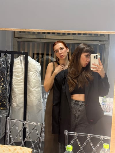 Two young women are taking a mirror selfie in what appears to be a dressing room or backstage area. The woman on the left has long red hair and is wearing a sleeveless brown dress and gold earrings. The woman on the right has long brown hair and is wearing a black blazer over a black crop top and grey jeans. She is holding a smartphone and taking the photo. In the background, there is a rack of clothes covered in plastic, and a clear acrylic chair. A bottle of water is visible on a ledge behind the woman on the right. The overall environment suggests a functional space, likely related to a performance or event. The city of Городской округ, Russia is not discernible from the visual information.