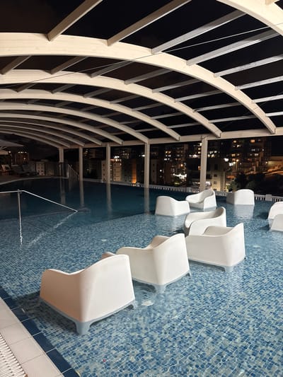 The image depicts a rooftop swimming pool area at night in Tirat Karmel, Israel. The pool is illuminated and filled with blue tiled water. Several white, modern-style lounge chairs are partially submerged in the water. A curved, white-paneled roof structure with black sheeting is visible overhead, supported by beams. In the background, city lights and buildings suggest an urban environment. A metal railing and steps are visible at the edge of the pool. There are no people present in the scene.