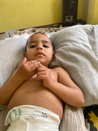 This image, captured in Dolni Tsibar, Bulgaria, features a young child, likely a toddler, lying on their back. The child has light brown skin, short dark hair, and dark eyes, looking directly at the viewer with a calm or neutral expression. They are shirtless and wearing a white disposable diaper with green and black patterns.

The child is resting on a bed or couch, supported by a light grey, textured pillow beneath their head and left arm. Another lighter, possibly striped pillow is partially visible to their right side (viewer's left). A blanket or fabric with a black and yellow/gold abstract design is seen behind the pillows. The background consists of a pale yellow wall, and in the upper left corner, a dark brown object, possibly furniture or an electronic device, with a small reflective component. The child's left hand is near their chin, and the right hand is closer to their chest. The indoor setting suggests daytime. No discernible text is visible within the image.