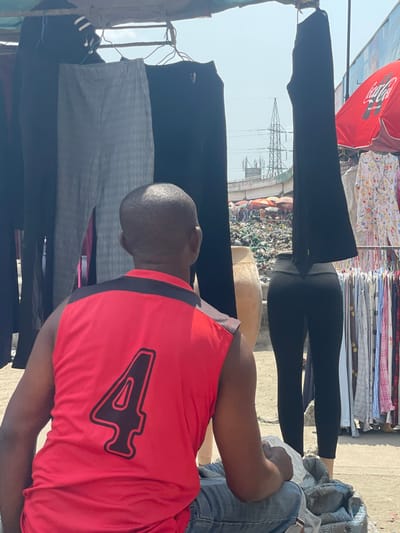 The image captures an outdoor market scene in Aiyetoto-Asogun, Nigeria, under bright daylight. A male individual, viewed from behind, is seated or crouching in the foreground, wearing a red sleeveless shirt with black shoulder accents and a large, black-outlined red numeral '4' on his back, along with blue jeans. His hands rest on a bundled item contained within a white plastic bag and a grey woven sack. Directly above and around him, a display of clothing hangs from a horizontal bar, including dark, grey plaid, and black trousers. To the right, a headless mannequin's lower body displays black leggings, adjacent to a partial view of a light-colored mannequin torso and a clothing rack with various garments, including a white floral dress. A red Coca-Cola branded umbrella is open on the right side of the frame. In the midground and background, an elevated concrete structure (likely an overpass) spans the scene, with a cluttered market area featuring various stalls and debris beneath it. A large metal electricity transmission tower is visible against a clear, bright sky behind the elevated structure. The ground appears to be uneven earth or pavement.