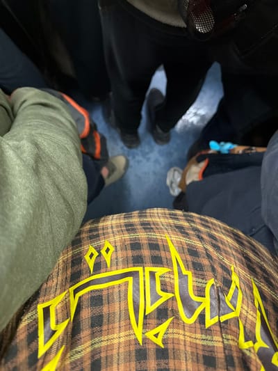 A bird's eye view of a crowded area with several pairs of shoes visible on a blue floor. A person in black pants and grey shoes stands to the left, near a bag. Another person wears tan shoes with orange detailing. An olive green garment covers the lower half of one leg. To the right, a brown and yellow plaid shirt dominates the frame, featuring a large, stylized yellow design.