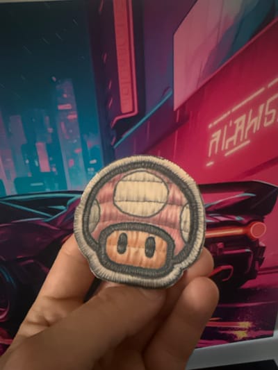 A close-up shot shows a hand holding a circular patch with a pink mushroom design, resembling a Super Mario mushroom. The patch has stitched details and a black outline. In the background, a vibrant, neon-lit cityscape is visible, with abstract buildings and a futuristic car. The overall aesthetic is reminiscent of cyberpunk or retro-futuristic art. The location is not specified.