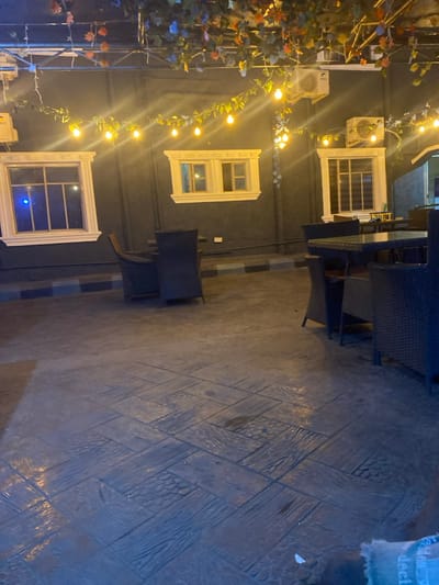 The image depicts an unoccupied outdoor commercial patio area at night. A dark-colored building wall serves as the background, featuring three white-framed windows with decorative lintels and sills. The leftmost window shows a blue light reflection from its interior. Two air conditioning condenser units are mounted on the wall above the central and right windows. Overhead and strung along the wall are garlands of artificial green and multi-colored foliage, interwoven with strings of warm yellow incandescent-style light bulbs. The ground is paved with patterned concrete, resembling a mosaic of wood planks and scale-like textures in grey tones. A low curb or border, striped in black and light blue/grey, runs along the base of the wall. Multiple dark-colored, wicker-style chairs are arranged around two square tables, one featuring a glass top. No individuals, actions, or interactions are present in the scene. The specific city of Igbessa, Nigeria, cannot be confirmed from visual cues within the image.