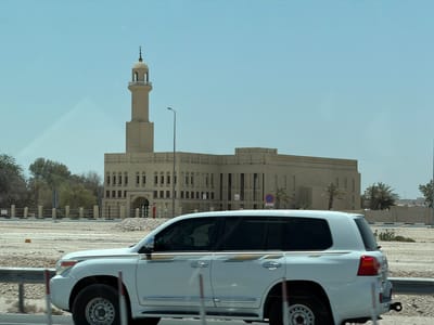 A white Toyota Land Cruiser SUV drives along a road in Umm Salal Ali, Qatar, under a clear, bright blue sky. The vehicle, observed from its left side, features tinted windows, gold and brown decorative stripes along its body, and a visible driver silhouette. Motion blur in the foreground suggests the car is moving.

In the background, a large, light-tan mosque dominates the scene. Its traditional architecture includes multiple arched windows and a prominent, tall minaret on the left. The minaret is topped with a distinctive finial featuring a crescent moon and star. A street lamp stands between the minaret and the main mosque structure.

The landscape is arid, with sandy and rocky ground bordering the road. Scattered green trees and several palm trees are visible, particularly near the mosque grounds. A rustic fence, constructed from posts and ropes, runs along the edge of the mosque property. A red circular "No Parking" sign with a diagonal line and Arabic script (likely "ممنوع" for forbidden) is visible on the mosque's side, near a palm tree. Below it is a smaller white rectangular sign with unreadable text. A metal guardrail runs alongside the road in the foreground. The bright sunlight indicates it is daytime, likely midday or early afternoon, with clear weather.