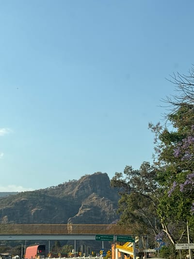 The image displays an exterior scene on a clear, sunny day, depicting a highway and mountainous landscape in Tepoztlán, Mexico. The upper two-thirds of the frame are occupied by a light blue sky. Below this, a range of rugged, dry mountains with sparse vegetation, including trees on ridges, extends across the mid-ground. Illumination indicates direct sunlight, casting shadows on the mountain slopes. In the lower portion of the image, a multi-lane highway system is visible. A yellow-barriered pedestrian overpass spans horizontally across the lower mid-ground. Below this, two green highway signs are present: one on the left reads "MEXICO Cuota CUERNAVACA Libre", and one on the right reads "TEPOZTLAN Libre". A partial red vehicle is visible on the far left lane, positioned below the overpass. On the far right, a tree with green foliage and purple flowers (Jacaranda) partially frames the scene, and a partially obscured white sign reads "PREPARE SU CUOTA AUTOS $". No individuals or specific actions are discernible. The scene appears to be captured from a vehicle on the highway approaching a toll plaza for Tepoztlán.