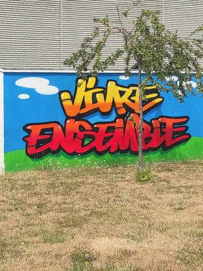 The image shows a mural with graffiti art on a blue wall. The graffiti reads "Vivre ensemble" in large, stylized lettering. The word "Vivre" is colored in yellow and orange gradients, while "ensemble" is in red and orange gradients. The letters have a 3D effect with black outlines. Above the text, there are two white cartoon-like clouds. The wall is bordered by a green painted stripe at the bottom, representing grass, with some white flowers. In front of the mural, a narrow tree with green leaves and small yellow fruits is growing. The ground in front of the wall is covered in dry grass and scattered white dandelion seeds. Behind the blue wall, there is a building facade with horizontal siding. The scene appears to be outdoors, possibly in a public area of Magny-le-Hongre, France, given the presence of graffiti art and an outdoor setting. There are no people visible in the image.