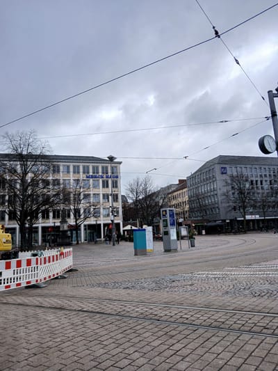 This photograph depicts a street scene in Darmstadt, Germany, characterized by overcast skies. The architecture includes multi-story buildings with repeating window patterns and a building with "MERCK HAUS" in blue lettering on its facade. Street infrastructure includes tram tracks curving across the paved surface, overhead tram lines, and streetlights with clocks. Barriers mark a construction or maintenance area in the foreground. People and vehicles are present but not prominent, adding to the urban context. The image captures a typical day in the city.