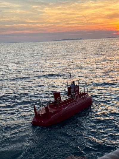 A small red submersible with a canopy is sailing on the sea near the coast of Primošten, Croatia. The submersible has a single occupant at the helm, facing away from the camera. The sun is setting, casting a warm glow over the sky and reflecting on the water. The sea is calm with gentle ripples. In the distance, faint outlines of islands can be seen. The submersible appears to be moving from left to right in the frame. The scene evokes a sense of tranquility and natural beauty.