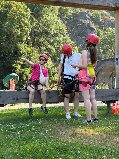 Three people, two women and one person of indeterminate gender, are standing and sitting on a wooden beam. The person sitting on the left, a woman wearing a pink t-shirt, red helmet, and sunglasses, points to the left with her right hand. She is wearing a harness and has a white sling on her left arm. The person standing in the middle, wearing a white t-shirt, black shorts, and a red helmet, is looking down at their phone. The woman standing on the right is wearing a light-colored tank top, pink shorts, and a yellow pouch on her back. She is wearing a harness. All three are wearing helmets and appear to be prepared for an outdoor activity, possibly zip-lining or a ropes course. They are in an outdoor setting with trees and a rocky incline in the background, suggesting a natural park or adventure site. Orange traffic cones are visible on the right side of the image. The location is identified as Warisy, Belgium.