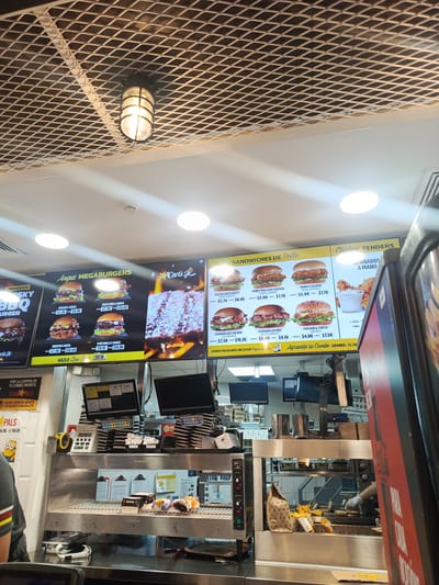 The image captures the interior of a Carl's Jr. fast-food restaurant in Samborondón, Ecuador. The scene is dominated by brightly lit digital menu boards showcasing various food items. On the left, "Angus MEGABURGERS" are advertised, including Original, Guacamole, Memphis, and Portobello Angus burgers, with prices ranging from $7.20 to $10.80 for combos. A "HAZLO Low Carh" option is also visible. To the right, "SANDWICHES DE Pollo" (Chicken Sandwiches) are displayed, featuring Bacon Swiss Crispy, Honey Mustard, Ranch, Chargrilled Club, Chargrilled BBQ, and Chicken & Cheese sandwiches, priced from $4.80 to $10.20. Further right, "Chicken TENDERS APANADOS A MANO" (Hand-Breaded Chicken Tenders) are shown. A sign states "COMBOS REGULARES INCLUYEN Papas y cola" (Regular Combos Include Fries and Cola) and "Agranda tu Combo GRANDE: $1.20" (Upgrade your Combo LARGE: $1.20).

Below the menu boards is the service counter and food preparation area. Cash registers and point-of-sale systems with numeric keypads are visible. A food warmer labeled "CAUTION HOT" holds boxed food items and fries. In the background, a person's gloved hand is seen near a grill, suggesting active food preparation. Bags of fries in branded cups are ready for serving. A sign for "STAR PALS" kids meals offers a choice of "UN JUGUETE, UN HELADO O UNA GALLETA" (a toy, an ice cream, or a cookie) with prices around $6.30. The ceiling features an exposed metal grid pattern with recessed lighting and a caged industrial-style light fixture. A fire alarm or smoke detector is also mounted on the ceiling.