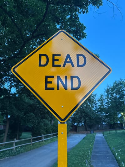A yellow diamond-shaped sign with black text reading "DEAD END" is mounted on a metal post on the right side of a paved path in Shaker Heights, United States of America. The sign is illuminated by ambient light, and its reflective surface is visible. Behind the sign, lush green trees form a dense canopy against a clear blue sky. To the left of the path, a rustic wooden fence runs parallel to a paved road. The path itself is bordered by grass and small, white-tipped garden lights. In the background, more trees and glimpses of houses suggest a residential area. The overall lighting indicates either early morning or late evening. There are no people or animals visible in the frame.