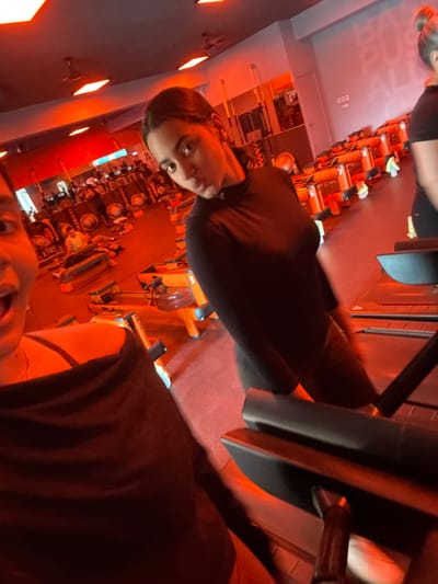The image depicts an indoor fitness studio, likely an Orangetheory Fitness facility, characterized by a predominantly red-orange ambient lighting scheme. This specific location is identified as Orangetheory Fitness at BlueMall in Santo Domingo, Dominican Republic, based on the signature aesthetic and partial text "BA... PUS... ALL..." visible on a rear wall, indicative of the brand's slogans.
The scene features three individuals within the gym environment. In the immediate foreground, a person (Subject 1) is partially visible on the left, capturing the photograph as a selfie. Only their left eye, cheek, and open mouth, suggesting an expression of surprise or excitement, along with a portion of a dark garment, are discernible.
In the midground, centrally positioned, stands a female (Subject 2) wearing a long-sleeved black athletic top and dark leggings. She has dark hair pulled back and is looking directly at the camera with a slight pout. She is standing on a treadmill platform.
Further in the background, to the right of Subject 2, a third female (Subject 3) is partially visible from the back, showing the top of her head with hair in a bun and the upper portion of a dark t-shirt. She is positioned on an adjacent treadmill.
The studio floor is dark, possibly a rubberized gym surface, intensely illuminated by the orange light. Numerous pieces of fitness equipment are visible throughout the space, including multiple treadmills, orange-framed WaterRower-style rowing machines (some bearing numbers like '3'), and aerobic steps. Overhead, recessed square light fixtures emit the pervasive orange glow, and ceiling fans are also present. The walls are a dark grey or black. The overall environment suggests an active or recently active fitness class setting.