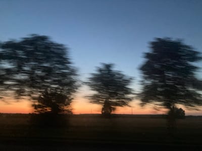 The image captures a blurry, impressionistic view of trees silhouetted against a twilight sky. The sky transitions from a pale blue at the top to a warm orange and pink near the horizon, indicating the time of day is either sunrise or sunset. The motion blur suggests the photo was taken from a moving vehicle. The trees are dark and appear in motion, their forms stretched horizontally across the frame. In the foreground, a dark, indistinct landscape is visible, possibly a field or a roadside. The overall mood is serene and contemplative, evoking a sense of fleeting moments and natural beauty. There is no visible text in the image. The location context is Ракітніцкі сельскі Савет, Belarus.
