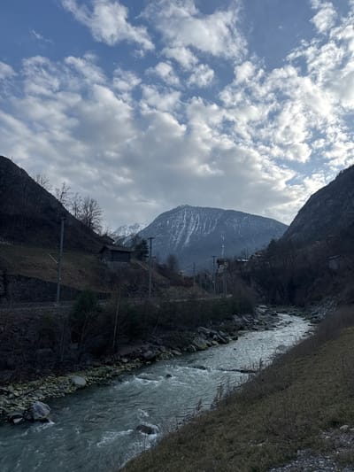 The image depicts a mountainous valley in Stalden (VS), Switzerland, observed from a slightly elevated perspective. A rapidly flowing, light green/turquoise river, consistent with the Matter Vispa, occupies the lower and central portion of the frame, winding from the lower left towards the mid-right. Its banks are rocky, with sparse vegetation.

On the left side of the river, a steep, dark slope ascends, featuring bare deciduous trees and some evergreen foliage. A single-track railway line, supported by a stone retaining wall in sections, traverses this slope, accompanied by multiple railway poles with overhead lines. A small, dark-timbered building with a brown roof is situated on this left slope, further back and above the railway line.

The right side of the river also presents a steep, grassy slope with sparse, brownish-green vegetation and a few bare bushes/trees.

In the mid-ground and background, multiple mountains are visible. A prominent, large mountain, partially covered in snow, dominates the center-to-right background. Other, higher snow-capped peaks are discernible in the far distance. Clusters of buildings are visible on the distant mountain slopes.

The sky overhead is partially covered by a mix of stratocumulus and altocumulus clouds, interspersed with areas of clear blue sky. The lighting suggests daytime, with diffused sunlight casting an even illumination across the scene. No individuals or animals are present.