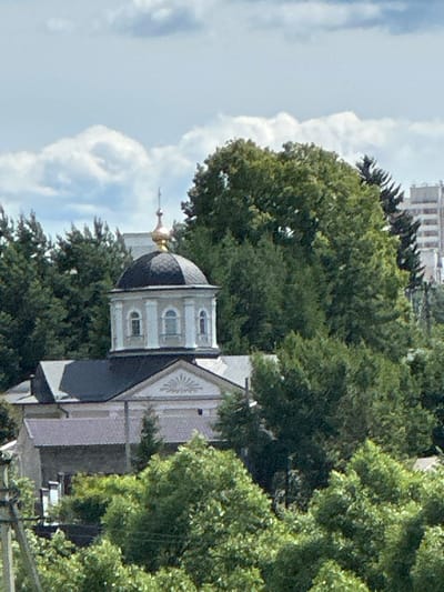 The image shows a view of the Church of the Nativity of the Blessed Virgin Mary in Dubrovitsy, Russia. The church features a prominent dark dome topped with a golden cross. The building has light-colored walls with arched windows and decorative elements. Surrounding the church is lush green foliage, with large trees obscuring parts of the structure and the sky. In the background, a multi-story building is partially visible, suggesting an urban or semi-urban setting. The sky is partly cloudy, with large white clouds against a light blue background. The lighting suggests it is daytime, likely mid-morning or afternoon, with bright sunlight illuminating the scene. There is no visible text in the image. The overall impression is a serene, almost majestic view of a historic religious building nestled within a natural environment.