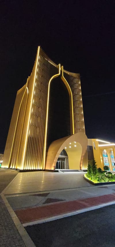 The image shows the exterior of a modern building at night, illuminated by warm golden lights. The building features a prominent, tall, arched entryway with vertical wooden slats and integrated lighting along its edges, giving it a sleek, contemporary appearance. The entryway leads to glass doors. To the right of the main structure, a lower, yellow-colored section of the building is visible, also with lighting accents. In front of the building, there is a paved walkway with geometric patterns and some low-lying greenery, including a small evergreen tree. The sky above is dark and clear. The building is identified as being located in the city of Satkak, Uzbekistan.