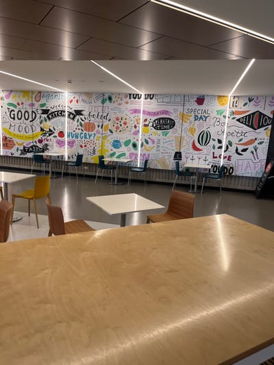 This media file captures an indoor dining or communal area, likely a cafeteria or food court, situated in Kelvin Grove, Australia. The image is taken at an angle, with a large, light-toned wooden table dominating the right foreground, its glossy surface reflecting ambient light.
The primary subject is a vibrant, floor-to-ceiling wall mural on the left, adorned with numerous hand-drawn illustrations of food items like carrots, watermelons, bananas, pizzas, avocados, pumpkins, and berries. Interspersed within these illustrations are various phrases and single words, alluding to food and a positive atmosphere. Linear LED lighting strips are visible along the ceiling and walls, providing illumination.
Several types of chairs are present around smaller white tables and along the larger wooden table. These include modern chairs with brown, leather-like upholstery, chairs with teal or dark blue seats, and at least one yellow chair. To the far left, a dark brown paneled wall, possibly made of wood, adds a contrasting texture. No people are visible, suggesting the area is empty or being photographed during an off-peak time.
Visible text on the mural includes: "DELICIOUS TO-GO", "Bon Appetit", "HAPPY", "SPECIAL DAY", "GOOD MORNING", "BREAKFAST TIME", "MEALS", "ADD BANANA SPIRULINA BERRIES", "LOVE", "LET'S GET BAKED", "HUNGRY?", "KITCHEN", "GOOD MOOD", "ORGANIC", "Healthy", "TASTE", "BRIGHT", and "CHIA". The overall impression is one of a modern, cheerful, and inviting space designed for enjoying food.
