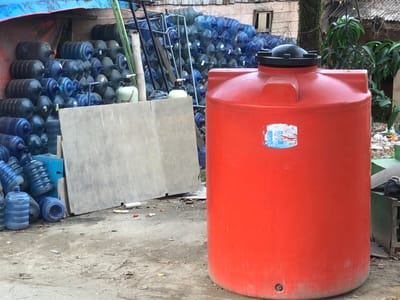 This is a daytime, eye-level, medium shot of a storage area in Pantegakibale, Indonesia. A large, bright orange water tank is in the foreground, occupying the right side of the frame. The tank has a black lid with handles and a small circular drain at its base. A sticker with some blue and red markings is affixed to the tank.
To the left of the tank, a large stack of blue and dark gray plastic water jugs is visible. These jugs are of the standard large capacity, often used for water dispensers. Behind the jugs, a tall, light gray, flat object, possibly a piece of plywood or concrete, leans against the wall.
Further in the background, a dense collection of blue and gray water jugs is organized in racks and stacked haphazardly under a blue tarp. Some bamboo poles and a metal frame are also part of the storage setup. The environment appears to be an outdoor lot, with loose dirt and debris on the ground. The buildings in the background seem to be made of concrete and corrugated metal. No people are visible in the image.