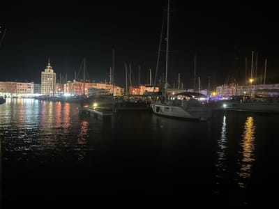 The image captures a nighttime view of the Agde marina in Agde, France, along the Hérault River. The scene is dominated by the reflections of colorful lights from buildings and boats on the dark, rippling water. A tall, illuminated building, likely a significant landmark in Agde, stands out in the background. Several sailboats are moored in the marina, their masts and rigging silhouetted against the night sky and the illuminated cityscape. The water is calm, with the lights creating long, shimmering reflections that stretch across the surface. The overall atmosphere is serene and picturesque, showcasing the beauty of the marina at night.