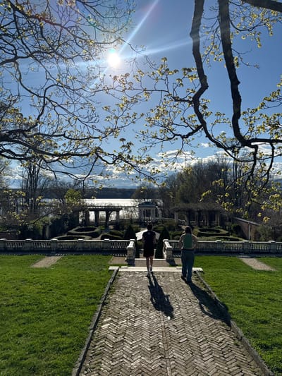 The image shows a scenic view in the Town of Red Hook, United States of America, featuring a garden overlooking a body of water, likely the Hudson River. Two people are standing on a herringbone brick path, casting long shadows. The garden is manicured with neatly trimmed hedges and grass. A decorative balustrade runs along the edge of the garden, defining the space. In the foreground, a tree with budding leaves frames the scene, partially obscuring the bright sun in the upper left corner. The sky is a clear blue with scattered clouds reflecting on the water's surface. In the background, trees line the opposite shore of the river.