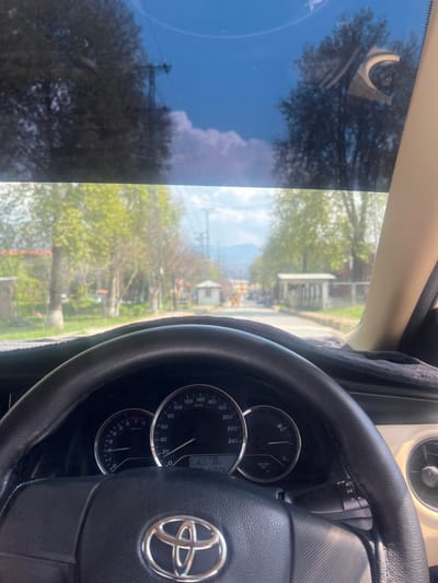 The image presents a forward-facing view from a stationary vehicle, revealing a tree-lined road in Abbottabad, Pakistan. The foreground is dominated by the vehicle's interior: a black, leather-wrapped Toyota steering wheel with a prominent silver logo, positioned directly ahead of a black dashboard covered by a textured black mat. Visible through the steering wheel are the instrument cluster dials, indicating zero speed (0 km/h) and zero RPM, with a digital display showing "13:25" and "41525" (likely odometer). The vehicle's interior trim on the right is beige/tan, with a circular air vent partially visible.
Through the windshield, which has a blue tint at the top, a straight paved road extends into the middle distance. Both sides of the road are lined with mature trees displaying vibrant, light green foliage, indicative of spring or early summer. Further down the road, two small, light-colored guard-hut-like structures are positioned on either side, suggesting a controlled entry or exit point. Beyond these structures, indistinct buildings are visible, and in the far background, hazy blue-grey mountains rise against a partially cloudy blue sky. Tall utility poles and overhead lines are present in the distance. The scene is brightly illuminated by daylight, with visible sun glare on the windshield. No clearly identifiable individuals or specific actions are discernible beyond the implicit presence of the person capturing the image from inside the vehicle. The overall environment suggests an urban or cantonment area typical of Abbottabad, characterized by its greenery and mountainous backdrop.