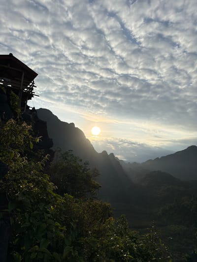 The image depicts a mountainous landscape with a structure built on a cliffside in the foreground. The sun is setting or rising, casting rays of light through the clouds and illuminating the distant mountain peaks. The overall scene is one of natural beauty and a remote location. The structure appears to be a small wooden building with a red roof, supported by stilts on the edge of the cliff. Lush green vegetation covers the cliff and a portion of the foreground. The mountains in the background are shrouded in mist and shadow, creating a dramatic and atmospheric view. The sky is filled with scattered clouds, some of which are illuminated by the sun, creating a textured and dynamic backdrop. The city of Naka, Laos, is not explicitly visible in the image, but the mountainous terrain and the style of the structure suggest a rural or natural setting consistent with such a location.