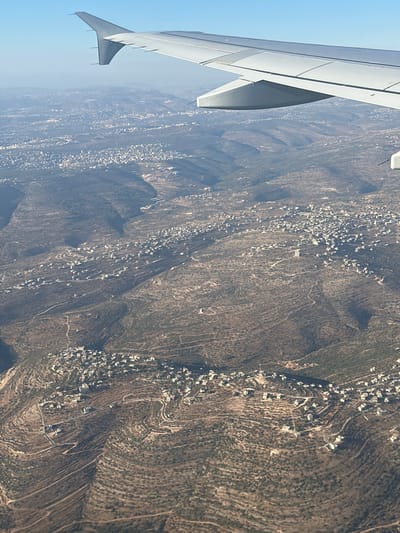 This is an aerial photograph, likely taken from an airplane, showing a view of a mountainous landscape dotted with buildings. The left side of the image is dominated by the wing of an aircraft, rendered in white with gray accents and visible winglets. The landscape below is a tapestry of green and brown, with evidence of terracing on the hillsides. Numerous white buildings are scattered across the slopes, appearing as clusters in some areas and more spread out in others. Winding roads or pathways connect these settlements, snaking their way through the terrain.
The lighting suggests it is daytime, with clear blue skies visible in the upper left corner. The overall impression is one of a rural or semi-rural settlement nestled within a challenging geographical environment. The perspective, looking down from above, emphasizes the scale of the terrain and the way human habitation has adapted to it. There are no visible people, specific objects other than the airplane wing and buildings, or any discernible text within the image. The location context provided is "Unknown, Palestinian Territories," which may hint at the style of architecture or the type of landscape, but this cannot be definitively confirmed from the image alone. The scene conveys a sense of quiet observation, looking down upon a community from a distance.
