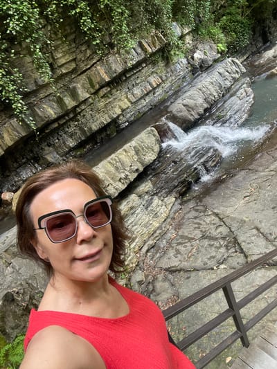 A woman takes a selfie in front of a waterfall in the city of Bolshoy Sochi, Russia. She is wearing a red top and large square sunglasses. The waterfall flows over layered gray rocks covered in green moss and vines. A wooden railing is visible in the lower right corner of the image.