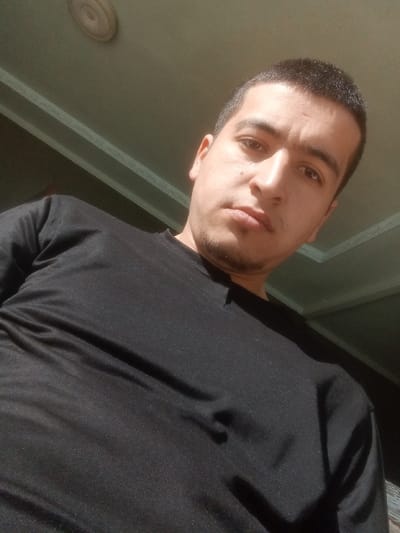 This image, originating from Kasan, Uzbekistan, features a single male individual. The subject is seen from a low-angle perspective, positioned indoors. He has short, dark hair, dark eyes, and light stubble on his chin. A small blemish is present on his right cheek (viewer's left). The man wears a plain black, long-sleeved shirt and gazes directly forward with a neutral expression. The background displays a light-colored ceiling with a circular light fixture visible in the upper left corner. A horizontal ceiling trim or architectural detail spans across the upper portion of the frame. Illumination appears to originate from the viewer's left, highlighting the subject's right side and creating subtle shadows on his left.