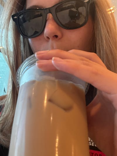 A woman with long blonde hair and black sunglasses is drinking from a tall plastic cup filled with a light brown liquid, likely coffee or tea. Her hand partially covers the cup. The image is shot from a slightly angled perspective, with the cup and her face in the foreground. The lighting appears to be indoors, and there is a glimpse of a sign on the cup and a tiled floor behind her.