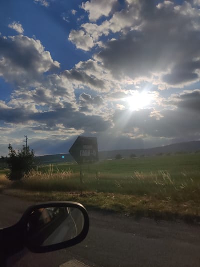 The image depicts a view from inside a vehicle, looking out towards a rural landscape under a dramatic sky. The foreground shows the side mirror of a car, reflecting an interior with what appears to be a person. Beyond the car's immediate presence, a road runs along the right side of the frame, bordered by tall grass and vegetation.

In the middle ground, a dark, hexagonal road sign stands prominently. The sign contains text, with "MEKAHЯ" visible in the upper section, and "ГАБИ-В" below it in larger Cyrillic letters. This sign indicates the direction to "Gabbi-V" and possibly a settlement or location named "Mekahya." Such signs are common in rural areas of Bulgaria, directing travelers to villages, farms, or specific points of interest.

The background reveals a gently rolling green field, extending towards a distant line of hills or mountains. The sun is intensely bright, positioned high in the sky and partially obscured by a mixture of dark and bright clouds, creating striking sunbeams that pierce through the atmosphere. The overall lighting suggests it is either late afternoon or early evening. The presence of a lone utility pole with wires near the sign suggests proximity to some form of infrastructure, though the landscape remains largely undeveloped. Given the Cyrillic script on the sign, the location is likely in Bulgaria, and the sign's content specifically suggests a connection to the village of Belanitsa, Bulgaria, a common practice for local signage.