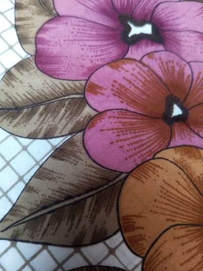 The image features a close-up view of a floral print fabric. Large flowers in varying shades of pink, coral, and orange dominate the scene. The petals display a gradient effect, transitioning from lighter to darker hues. Brown leaves with distinct venation are interspersed among the blossoms. A white background is visible, adorned with a subtle grid pattern in a light brown color. The overall impression is a warm, vintage-inspired textile design.