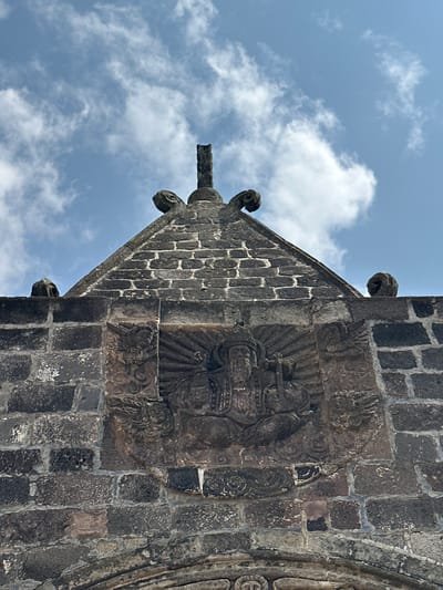 This is a low-angle, medium shot of the facade of the San Andrés Apóstol Church in San Andrés Calpan, Mexico. The building is constructed of dark, rough-hewn stone blocks. The top of the image is dominated by a blue sky with scattered white clouds.

Centered on the facade, below a triangular pediment, is a stone relief carving. The carving is oval in shape and depicts a central figure radiating light, possibly a saint or deity. The figure has a long beard, wears a crown or elaborate headdress, and holds an object in its hands, possibly a scepter or staff. Flanking the central figure are smaller, winged figures with cherubic faces, and swirling cloud or floral motifs. The relief is weathered and worn, with some details obscured by time and the elements.

Above the relief, the pediment is made of smaller, stacked stone blocks, and is topped with a dark stone cross. On either side of the pediment, near the top corners of the stone structure, are small, carved gargoyle-like figures. The overall impression is one of historical and religious significance.