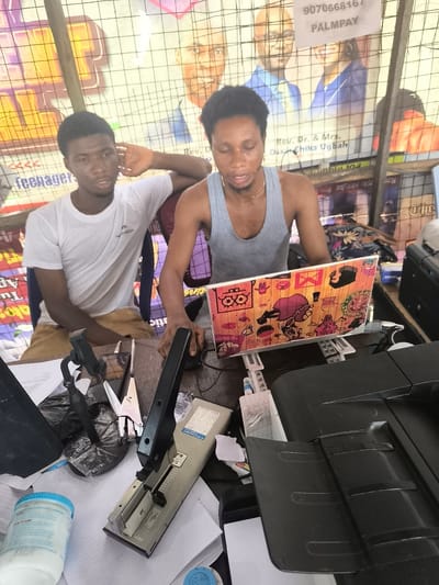 The image depicts an indoor or covered workspace, possibly a small business center, in Abraka, Nigeria, featuring two young men. The background shows several posters visible through a wire mesh fence.
On the left, a young man with dark skin and short dark hair is seated, wearing a white t-shirt with a "MXMECH" logo and light brown shorts. He appears to be observing, with his left arm resting on his chair and hand near his ear. On the right, another young man, also with dark skin and short dark hair, wears a grey tank top and a gold chain. He is actively using a laptop and an external mouse, looking focused.
The desk in the foreground is cluttered with various office supplies. The laptop lid, facing the viewer, is uniquely decorated with colorful, cartoon-like stickers. To the left of the laptop are a large, beige-colored stapler (with "EASY CLEARING JAMMED STAPLES" text), a mobile phone mounted in a holder, and a smaller, silver stapler. A white plastic container with a blue lid, labeled "HOW TO USE," is also visible on the desk. A black printer is positioned on the right side of the desk.
Visible text on the background posters includes a phone number "907066816" with "PALMPAY" below it, indicating a mobile payment service. Other text mentions "Rev. Dr. & Mrs. Oss Chika Ugbah" and partially visible words like "teenager." The scene captures a typical local business environment.