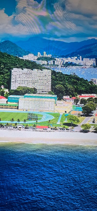 The image depicts an aerial view of a coastal city with mountains in the background and the ocean in the foreground. The city features a dense collection of high-rise buildings nestled on hillsides covered in lush green vegetation. In the mid-ground, there are several sports facilities, including a track and field stadium with a green field and distinct lanes, a swimming pool with blue tiling, and a tennis court. Adjacent to the sports complex are some smaller buildings with red roofs. A sandy beach separates the sports facilities from the deep blue ocean, which is dotted with several white sailboats. The sky above is a mix of blue and white clouds, with hints of color suggesting a rainbow. The image quality is pixelated, indicating it may be a digital photograph or a screenshot. There are no people visible in the image. The overall scene conveys a sense of a modern city integrated with its natural environment. The prompt incorrectly places this scene in Lippstadt, Germany, which is an inland city and does not have a coastline or mountains like those depicted.
