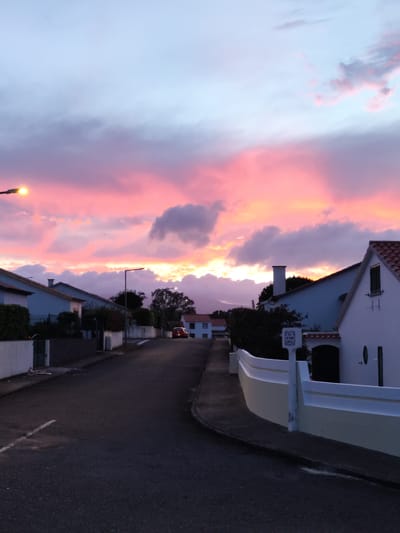 This is a vertical, eye-level, outdoor shot of a street at dusk in Pico da Pedra, Portugal. The sky is filled with dramatic pink and purple clouds, illuminated by the setting sun. On the left side of the frame, a streetlamp with a glowing yellow light is visible. The street is paved with asphalt and has a white dashed line marking the center. On either side of the street are houses with white walls and tiled roofs, typical of Portuguese architecture. Some houses have manicured hedges and trees. A red car is parked on the right side of the street, further down. The scene conveys a peaceful and serene atmosphere of a quiet residential street at the end of the day.