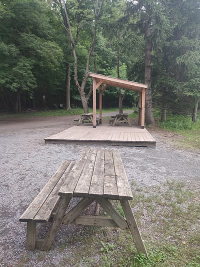 The image shows a park setting in Saint-Bruno-de-Montarville, Canada. In the foreground, a weathered wooden picnic table is positioned on a gravel surface, with grass and foliage bordering it. Behind this table, a wooden deck has been constructed, serving as a platform for another picnic table situated beneath a simple wooden shelter. This shelter has a slanted roof supported by wooden posts. The entire scene is surrounded by a dense forest with tall trees, suggesting an outdoor recreational area. A dirt path winds through the trees in the background. The lighting suggests it is daytime, likely overcast, as there are no strong shadows. The atmosphere appears peaceful and natural, with the wooden structures blending into the environment. No people are visible, and there is no visible text.