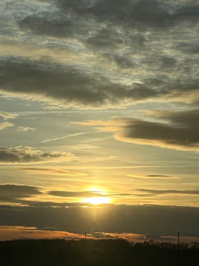 A landscape image shows a vibrant sunset. The sun, a radiant golden orb, dominates the center, casting a warm glow across the sky. Wispy clouds, painted in shades of grey and gold, stretch vertically. A dark, silhouetted horizon line runs along the right edge, punctuated by the stark vertical lines of utility poles. The sky is the dominant element, filled with dynamic cloud formations.