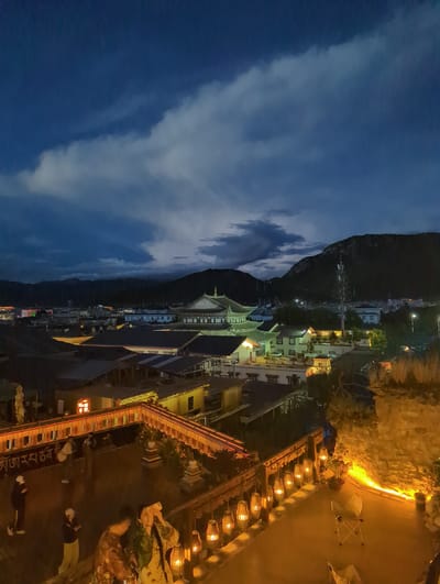 This is an aerial, nighttime shot of a village nestled at the base of a dark, imposing mountain range. The sky is a deep blue with scattered, wispy clouds that are illuminated by an unseen light source. The village is lit up with numerous lights, suggesting it is inhabited and active.

The most prominent structure is a building with a distinct, ornate roofline, likely a temple or monastery, given the location context of Shangri-La, China. This building is illuminated with warm, yellow light, and its architectural style is distinctly East Asian. Surrounding it are other buildings of varying heights and designs, some appearing more modern with multiple stories and windows, while others are lower and more traditional.

In the foreground, there is a brightly lit walkway lined with rows of decorative lamps. The warm glow of these lamps illuminates the faces of several people gathered on this walkway, some looking towards the village, others engaged in conversation. One person is holding up a smartphone, possibly taking a picture or video. The overall mood is one of quiet evening activity, with a sense of wonder and perhaps a touch of spirituality evoked by the illuminated temple and the dramatic mountain backdrop. The time of day is clearly night, and the weather appears clear, judging by the visible stars and the distinctness of the clouds. A banner with Tibetan script is visible near the bottom of the frame.