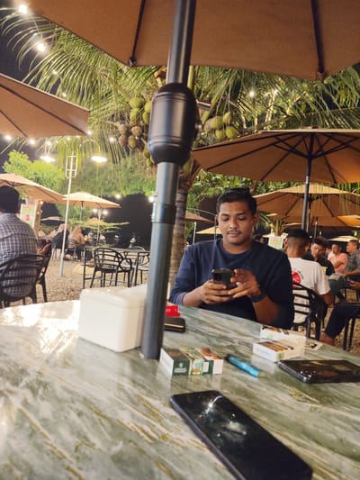 The image depicts an outdoor seating area at night, likely a restaurant or cafe, in Glokaron, Indonesia. The foreground features a marble-patterned table with several objects, including two mobile phones, cigarette packs, and a white tissue box. In the mid-ground, a man is seated at a table, looking at his phone. He is wearing a dark blue long-sleeved shirt. Behind him, other patrons are visible, seated at tables under large tan umbrellas. Palm trees with coconuts are in the background, illuminated by string lights, contributing to a tropical ambiance. The ground appears to be covered in gravel. The overall scene suggests a relaxed evening gathering.