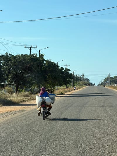 A person rides a motorcycle on a paved road. The motorcycle is heavily loaded with bags and containers. The rider wears a dark-colored head covering and a blue shirt. The road runs straight into the distance, lined with utility poles and trees. The sky is clear and blue. This photo was taken in the Muxara area of Mozambique, a small municipality.