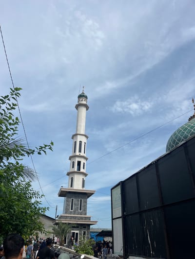 A tall minaret of a mosque towers against a partly cloudy sky in Paya Geli, Indonesia. The minaret is white with alternating sections containing arched windows and gray sections. A small green dome tops the minaret. Black roof-like structures overhang the right side of the frame, partially obscuring the building attached to the minaret. A gathering of people and parked motorbikes fill the lower right foreground of the image. Green foliage occupies the lower part of the frame. Electrical wires crisscross the sky between the minaret and the foliage.